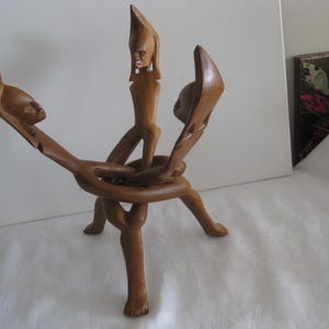 May include: A wooden sculpture featuring three stylized human figures intertwined. The figures are carved from light brown wood and connected by a circular base. The sculpture stands on three legs and has a natural finish.