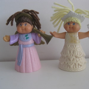 May include: Two vintage Cabbage Patch Kids angel dolls. One doll wears a pink dress and plays a horn. The other doll wears a white dress and holds a snowflake. Both dolls have blue eyes and yarn hair.