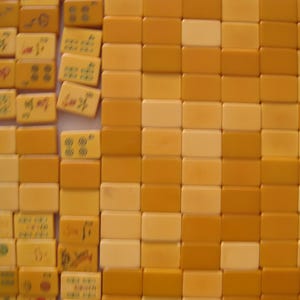 May include: A collection of rectangular mahjong tiles arranged in a grid. The tiles are primarily shades of yellow and cream. Some tiles feature engraved symbols, characters, and circular patterns in red, green, and black, while others are blank.