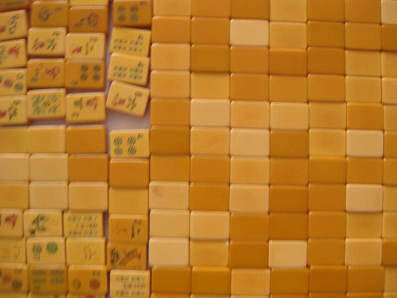 May include: A collection of rectangular mahjong tiles arranged in a grid. The tiles are primarily shades of yellow and cream. Some tiles feature engraved symbols, characters, and circular patterns in red, green, and black, while others are blank.