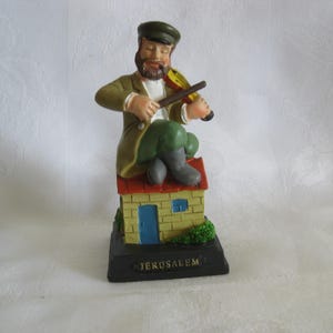 May include: A painted figurine of a bearded person wearing a green cap, brown jacket, and green trousers, sitting on a yellow brick house with a red roof while playing a yellow violin. The base is black with the word JERUSALEM printed in gold.