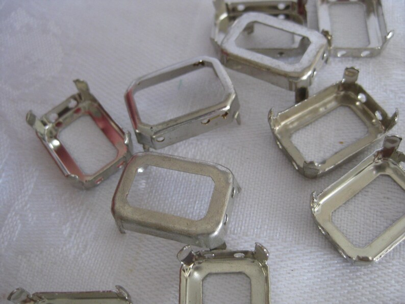 May include: A collection of silver-toned metal rectangular settings for jewellery making. Each piece features a hollow centre with four small prongs extending from the corners, designed to hold a stone or cabochon securely in place.
