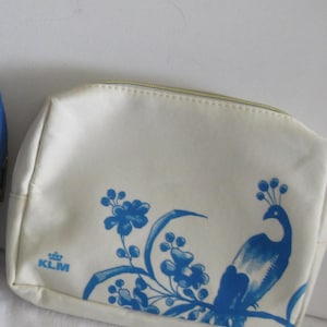 May include: Off-white cosmetic bag with a blue peacock and floral design. The bag has a gold zipper and the KLM logo. The peacock is perched on a branch with flowers.