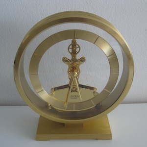 May include: A gold-colored quartz clock with a unique design. The clock features a circular frame with two concentric rings and a central decorative element. The base is rectangular, and the clock face is visible. The clock is labeled "Linden Quartz".