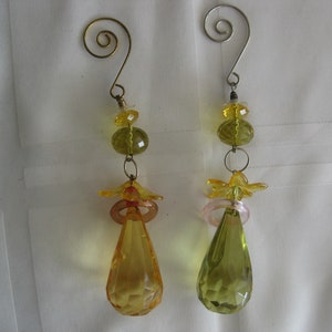 May include: Two decorative hanging ornaments with a spiral top. Each ornament features a series of yellow and green beads, a flower-shaped accent, and a faceted teardrop-shaped crystal. The ornaments are suspended against a white background.