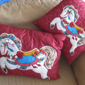 May include: Two red decorative pillows with a quilted pattern. Each pillow features a white carousel horse applique with floral accents, a blue saddle blanket, and a red bow. The pillows are on a beige sofa.