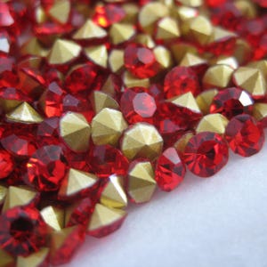 May include: Close-up of a collection of red and gold rhinestones. The red rhinestones are faceted and round, while the gold ones are pyramid-shaped. The gems are scattered across a white surface, creating a sparkling effect.