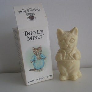 May include: A white cat-shaped figurine stands next to a box. The box is labeled "Toto Le Minet" and features a cartoon cat wearing a blue outfit. The box also has the Crabtree & Evelyn London logo.