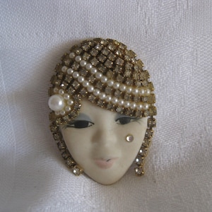May include: Oval-shaped vintage brooch featuring a stylized face. The face is adorned with a headdress of rhinestones and pearls. A single pearl accents the side, and a rhinestone adds a touch of sparkle to the cheek. The brooch has a gold-tone metal frame.