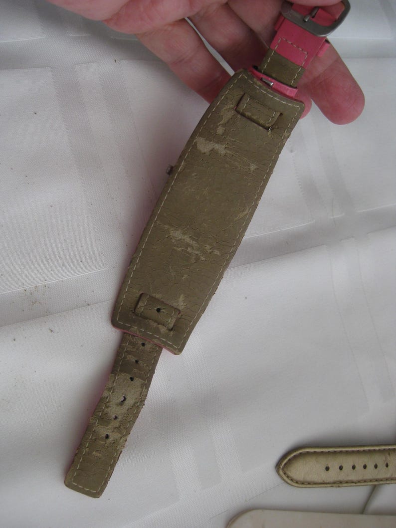 K&ouml;nnte beinhalten: Ein braunes Lederarmband mit einem rosafarbenen Lederband und einer silbernen Schlie&szlig;e. Das Armband hat mehrere L&ouml;cher zum Einstellen der Gr&ouml;&szlig;e.