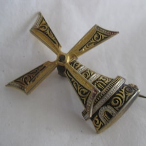 May include: A vintage windmill brooch with gold-tone metal, black enamel, and silver accents. The pin features a detailed design of a windmill with four blades and an ornate body. The brooch has a pin closure on the back.