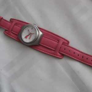 K&ouml;nnte beinhalten: Eine Armbanduhr mit rosafarbenem Lederarmband, wei&szlig;em Zifferblatt und silbernen Akzenten. Das Zifferblatt der Uhr zeigt das Puma-Logo und das Wort "Puma" in Rot.