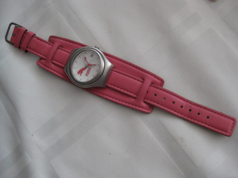 K&ouml;nnte beinhalten: Eine Armbanduhr mit rosafarbenem Lederarmband, wei&szlig;em Zifferblatt und silbernen Akzenten. Das Zifferblatt der Uhr zeigt das Puma-Logo und das Wort "Puma" in Rot.