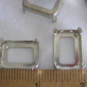 May include: Silver-toned metal rectangular prong settings for jewellery making. The settings feature four upright prongs at the corners and are positioned next to a wooden ruler showing a measurement of approximately 3.8 cm in width.