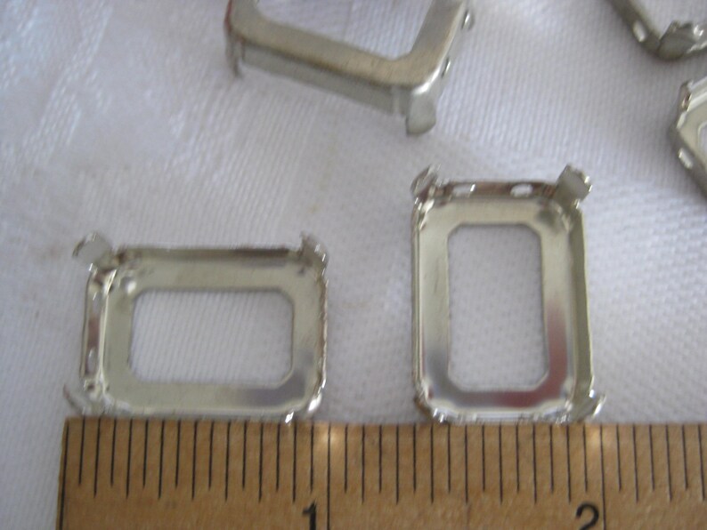 May include: Silver-toned metal rectangular prong settings for jewellery making. The settings feature four upright prongs at the corners and are positioned next to a wooden ruler showing a measurement of approximately 3.8 cm in width.