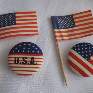 May include: Two small American flags on wooden sticks with a red, white, and blue button that says "U.S.A."