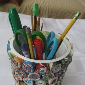 May include: A colorful pencil holder made from rolled up magazine pages. The holder is filled with various writing utensils, including pencils, pens, markers, scissors, and a green eraser.