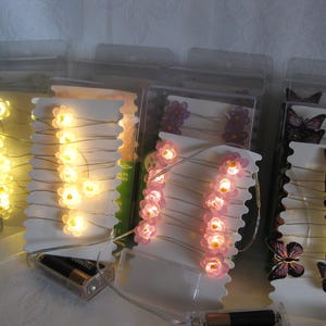 May include: Several strings of decorative lights are displayed, each featuring small, illuminated flower or butterfly shapes. The lights are packaged in clear plastic containers with white backing cards. The flower lights glow yellow and pink, while the butterfly lights are purple.