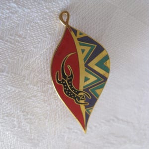 May include: A gold-toned pendant with a leaf shape. The pendant is divided into two sections: one red with a black jaguar design, and the other with a geometric pattern in green, blue, and gold. The pendant has a loop at the top.