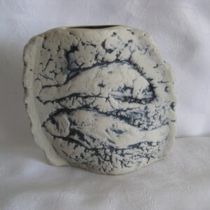 May include: A cream-colored ceramic vase with a textured, uneven surface featuring a relief design of two fish stacked horizontally. The recessed areas of the textured pattern are highlighted with dark blue or black glaze, creating a high-contrast look.