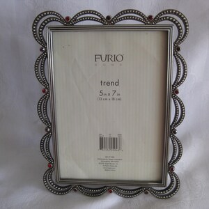 May include: A silver-toned metal picture frame with a decorative, scalloped edge. The frame has red accents and holds a white insert. The text "trend" and dimensions 5 in x 7 in are visible.