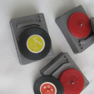 May include: Miniature gray record players with black and red vinyl records. One record has a yellow label with "Capitol" text, and another has a red label with "M-G-M" text. The records are on a white surface.