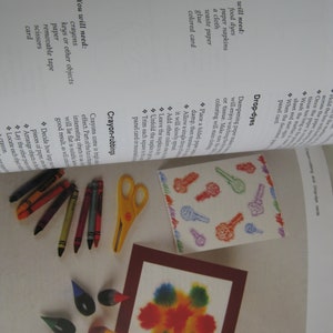 May include: An open book page with art supplies. Crayons, scissors, and paint drops are arranged near a framed watercolor painting. A key-patterned design and instructions are also visible.