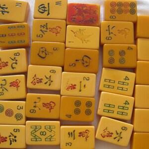 May include: A collection of rectangular yellow mahjong tiles arranged in rows. The tiles feature various engraved symbols, Chinese characters, and numbers in red, green, and black ink. Some tiles are blank, while others display intricate designs.