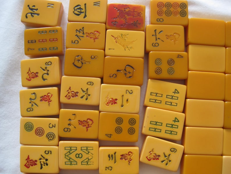 May include: A collection of rectangular yellow mahjong tiles arranged in rows. The tiles feature various engraved symbols, Chinese characters, and numbers in red, green, and black ink. Some tiles are blank, while others display intricate designs.