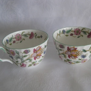 May include: Two white teacups with floral patterns. The cups feature a delicate design of pink, yellow, and blue flowers with green accents. Each cup has a curved handle and a green trim along the rim.