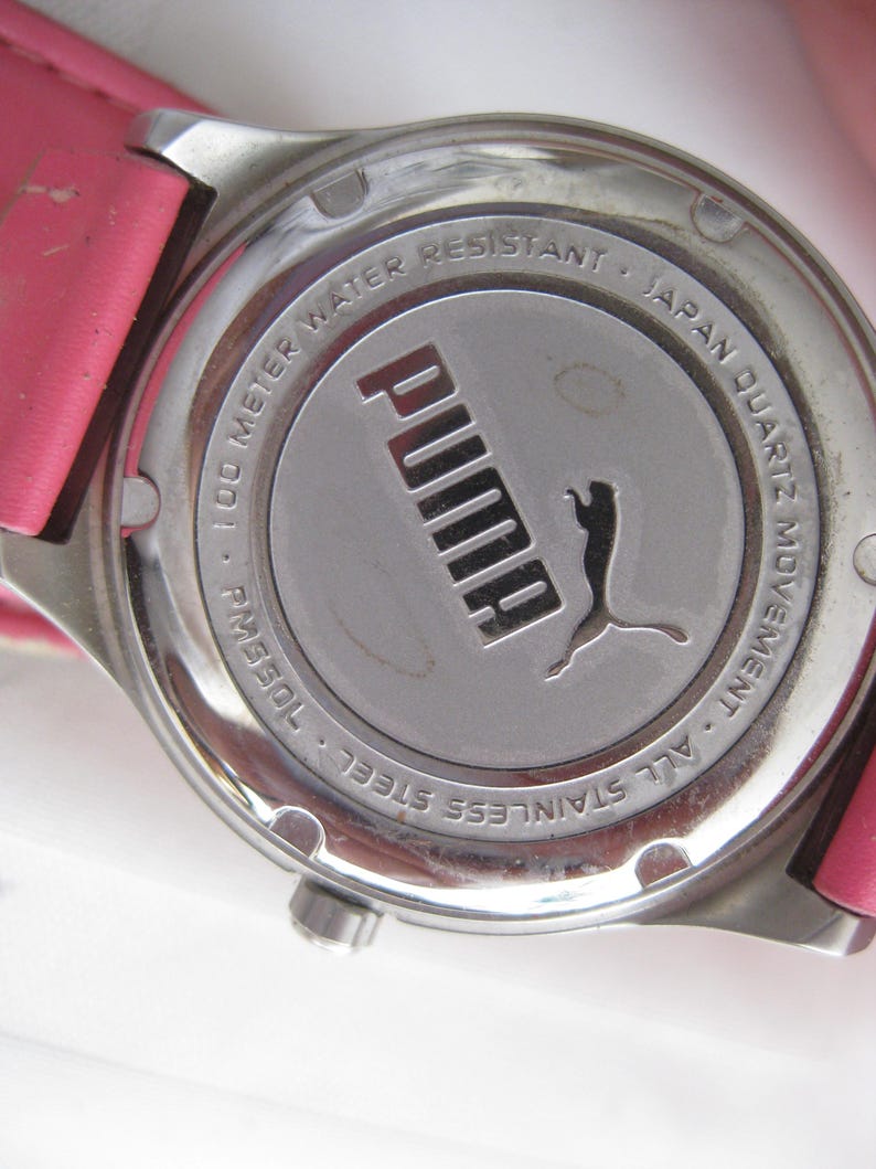 K&ouml;nnte beinhalten: Eine silberfarbene Uhr mit einem rosafarbenen Lederarmband. Das Zifferblatt der Uhr ist von einer silbernen L&uuml;nette mit dem Text "100 Meter wasserdicht - Japanisches Quarzwerk - Ganz aus Edelstahl" und dem Puma-Logo umgeben.