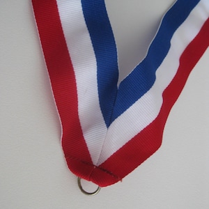 May include: A close-up of a red, white, and blue ribbon, likely for a medal or award. The ribbon is folded, with a metal ring at the bottom. The colors are vibrant and the ribbon appears to be made of a sturdy material.