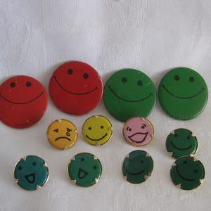 May include: A collection of colorful smiley face buttons and pins. The buttons are red, green, yellow, and pink, with black eyes and smiles. Some pins have gold-colored edges. The expressions vary from happy to sad.