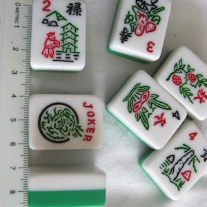 K&ouml;nnte beinhalten: Eine Sammlung wei&szlig;-gr&uuml;ner Mahjong-Steine. Die Steine zeigen verschiedene Symbole, Zahlen und chinesische Schriftzeichen in Rot und Schwarz. Ein Stein ist mit "JOKER" markiert. Ein Lineal liegt neben den Steinen und zeigt metrische Ma&szlig;e.