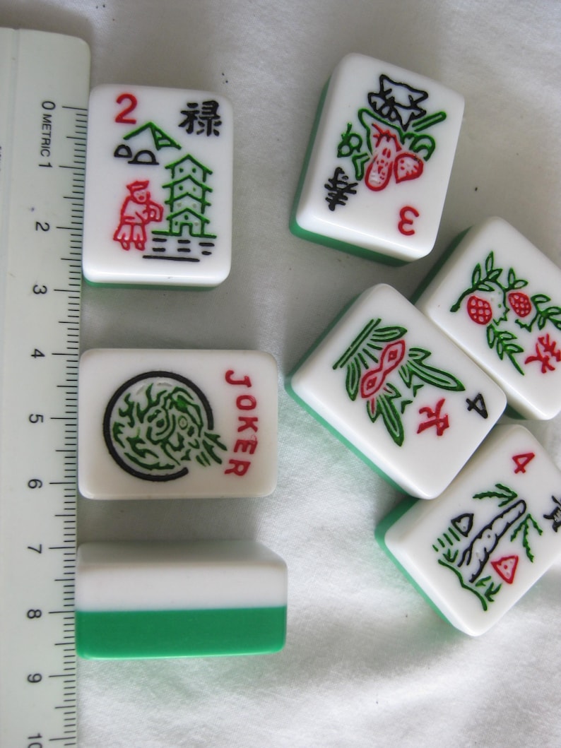 K&ouml;nnte beinhalten: Eine Sammlung wei&szlig;-gr&uuml;ner Mahjong-Steine. Die Steine zeigen verschiedene Symbole, Zahlen und chinesische Schriftzeichen in Rot und Schwarz. Ein Stein ist mit "JOKER" markiert. Ein Lineal liegt neben den Steinen und zeigt metrische Ma&szlig;e.