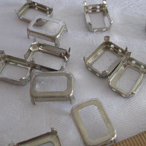 May include: A collection of silver-toned metal rectangular prong settings for jewelry making. Each setting features four small prongs at the corners and a hollow center. A wooden ruler is partially visible in the corner for scale reference.