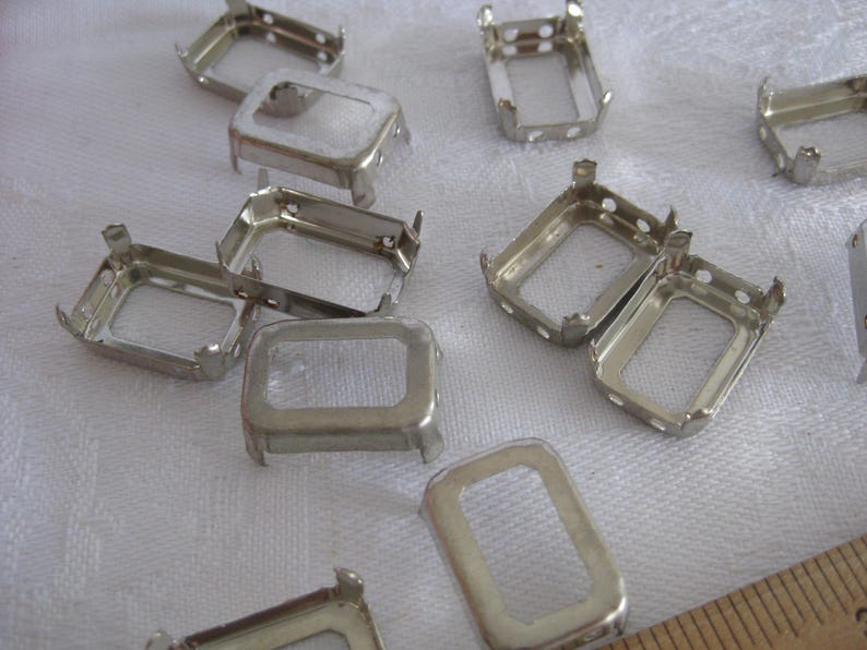 May include: A collection of silver-toned metal rectangular prong settings for jewellery making. Each setting features four small prongs at the corners and a hollow centre. A wooden ruler is partially visible in the corner for scale reference.