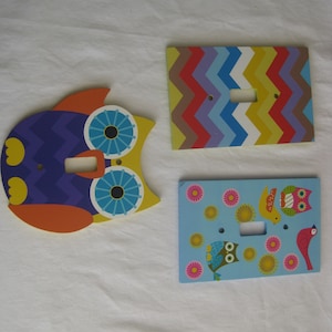May include: Three decorative wooden light switch covers. One is shaped like an owl with purple, orange, and yellow details. Another has a colorful chevron pattern. The third features birds and flowers on a blue background.