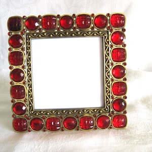 May include: Square picture frame with a brass-colored metal border. The frame is adorned with large, faceted red jewels and smaller red gems. The frame has a white interior.