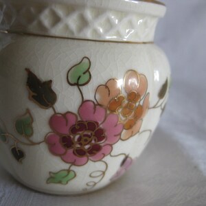 May include: A small, off-white ceramic jar with a floral design. The jar features pink, orange, and brown flowers with green leaves and gold accents. The lid has a diamond pattern. The jar has a crackle finish.