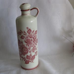 May include: A tall, off-white ceramic bottle with a handle and a stopper. The bottle is decorated with a floral pattern in shades of red. The design covers the majority of the bottle's surface.