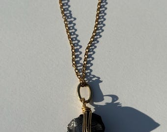 May include: A gold-colored chain necklace with a dark gray stone pendant. The stone is wrapped with gold-colored wire and hangs from a gold-colored oval loop. The necklace is displayed on a white surface.