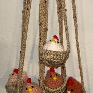 May include: Hanging crochet chicken decor featuring five chickens in woven baskets. The chickens are in various shades of pink, brown, and white, with red combs and yellow beaks. The hanging cords are made of a speckled brown yarn.