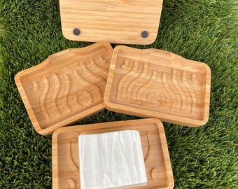 May include: A set of four bamboo soap dishes. Three dishes have a wavy design and one holds a white bar of soap. The dishes are rectangular with a curved handle. The fourth dish is a solid rectangle with four black rubber feet.
