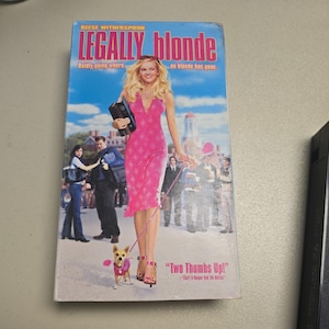 Puede incluir: Una cinta VHS de la película "Legally Blonde" con Reese Witherspoon. La portada muestra a Witherspoon con un vestido rosa, sosteniendo un maletín, con un perro pequeño. El título está en rosa y azul, con la frase "Boldly going where... no blonde has gone."