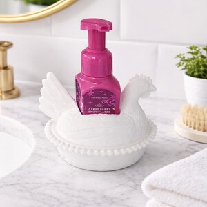 以下が含まれることがあります： ピンク色の石鹸ボトルが入った、白いセラミック製の鶏の形をした石鹸ディスペンサーホルダー。石鹸ボトルには「Strawberry Snowflakes」の文字が入っています。ホルダーは、背景に金の鏡がある大理石のカウンタートップにあります。