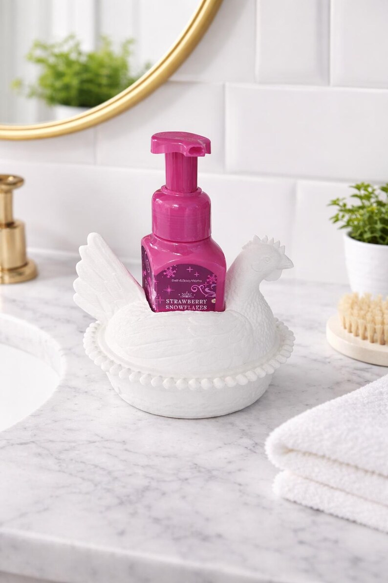 以下が含まれることがあります： ピンク色の石鹸ボトルが入った、白いセラミック製の鶏の形をした石鹸ディスペンサーホルダー。石鹸ボトルには「Strawberry Snowflakes」の文字が入っています。ホルダーは、背景に金の鏡がある大理石のカウンタートップにあります。