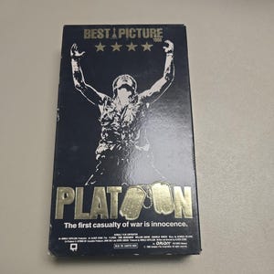 Könnte beinhalten: Eine VHS-Kassette für den Film "Platoon" mit dunkelblauem Cover. Das Cover zeigt eine Silhouette eines Soldaten mit erhobenen Armen, den Titel in goldener Schrift und den Text "Best Picture 1986".
