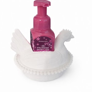 以下が含まれることがあります： ピンク色のハンドソープボトルとポンプトップが、白いセラミック製の鶏の形をしたホルダーに入っています。「Strawberry Snowflakes」と書かれたラベルが付いています。ホルダーは、翼と頭が見える鶏の詳細なデザインです。