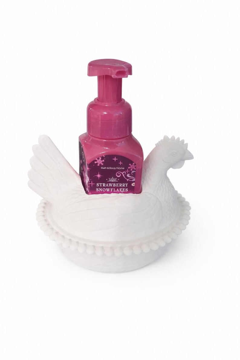 以下が含まれることがあります： ピンク色のハンドソープボトルとポンプトップが、白いセラミック製の鶏の形をしたホルダーに入っています。「Strawberry Snowflakes」と書かれたラベルが付いています。ホルダーは、翼と頭が見える鶏の詳細なデザインです。